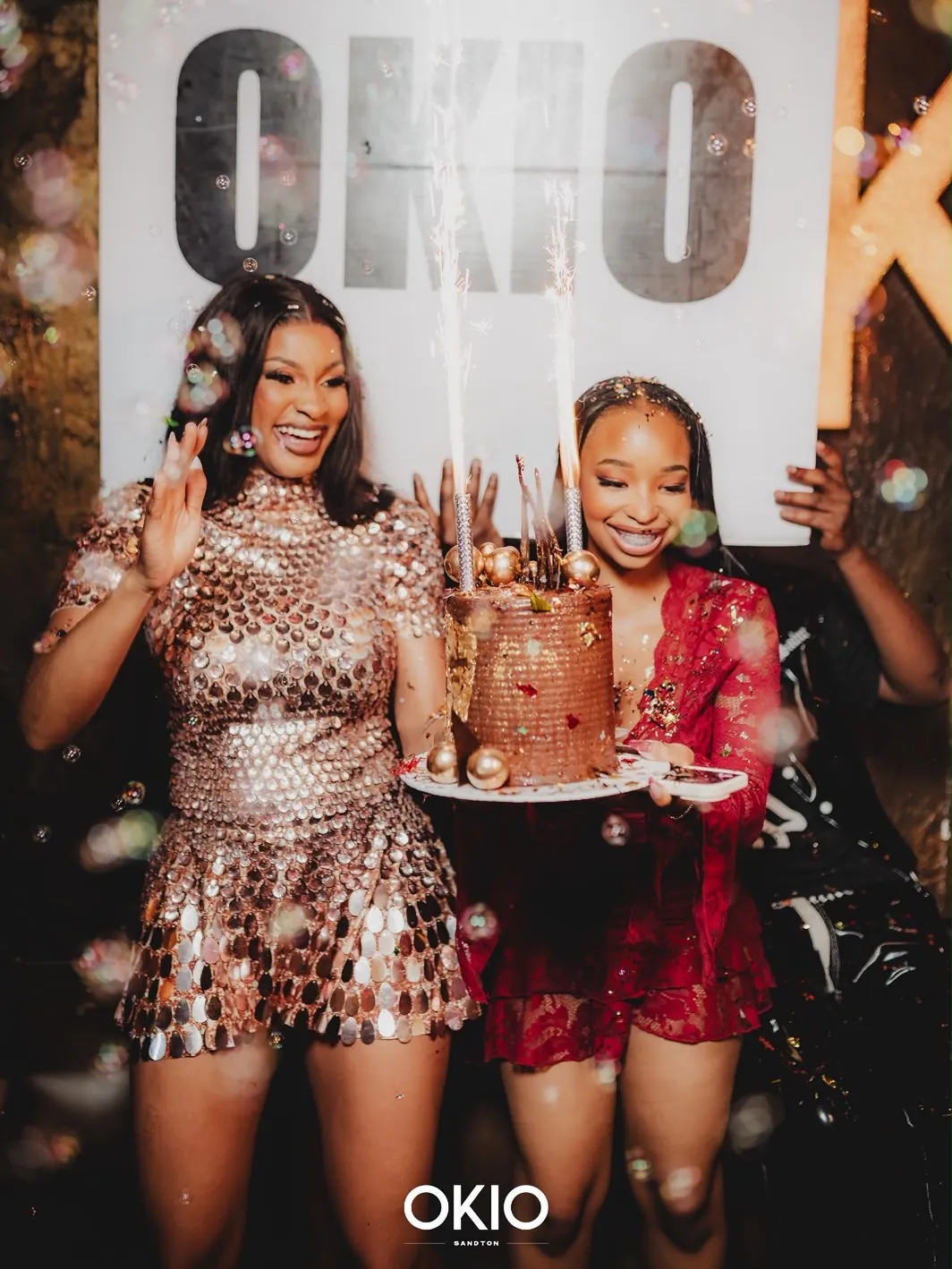 Birthday celebration with a gold-dressed guest and a sparkler cake in front of the OKIO wall